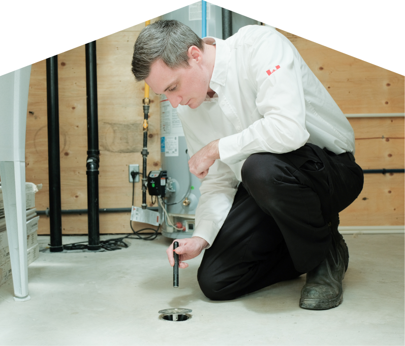 Dunn plumbing specialist inspecting a basement floor drain with a flashlight during a drain cleaning service, in a utility room with visible pipes, water heater, and wood panel walls.